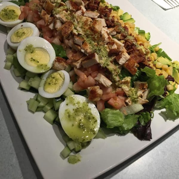 A large Cobb salad with chicken, eggs, tomatoes, avocado, corn, and greens on a white platter.